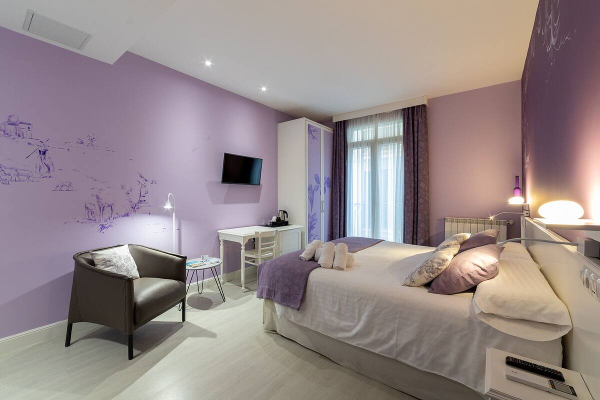 A bedroom with a bed, a desk, a chair, a TV, and a window with curtains.
