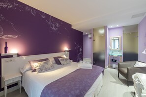 A modern bedroom with a large bed, a bedside table, a chair, a bathroom with a mirror and sink, and a wall with floral wallpaper. - Ur-Alde (San Sebastián)