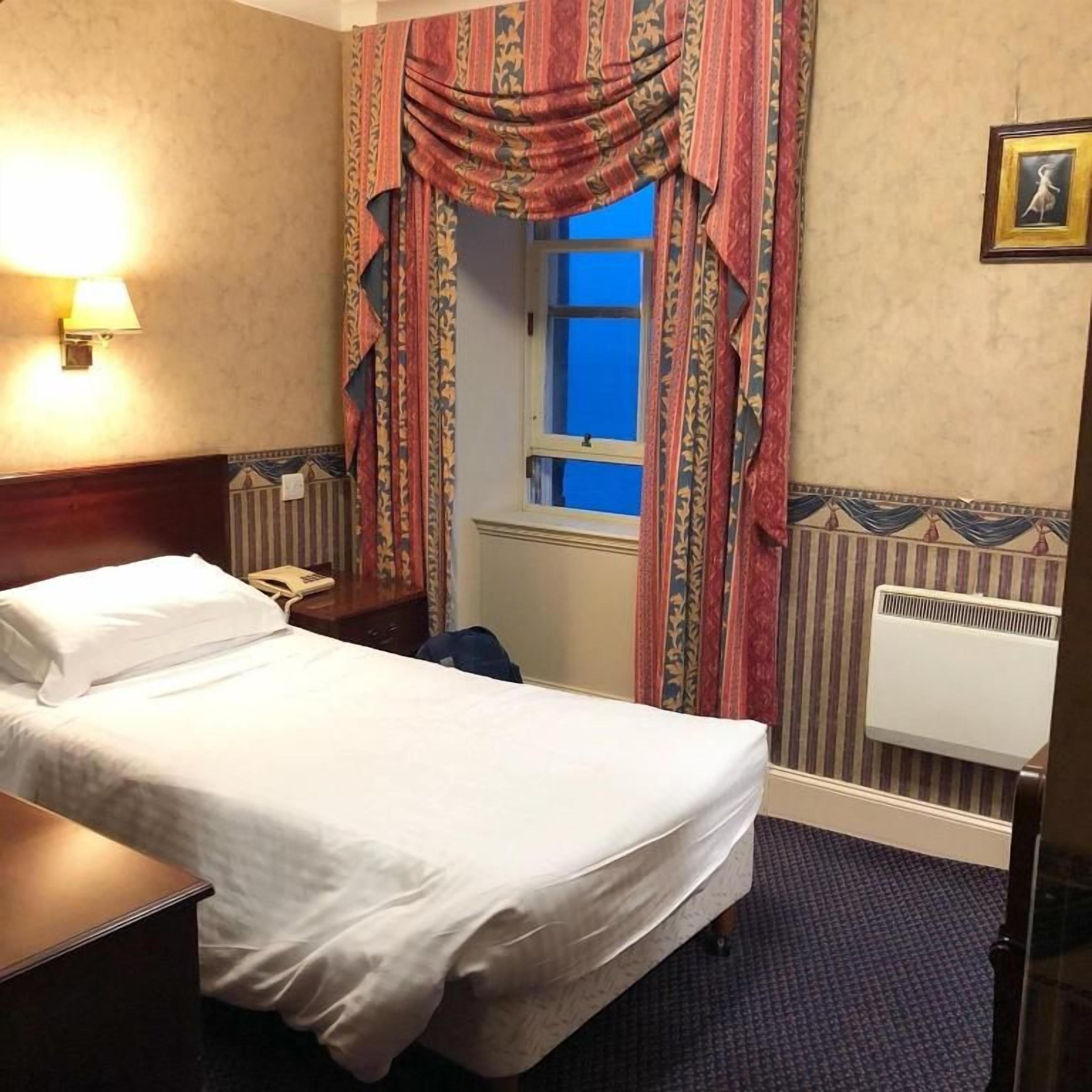 Una camera d'albergo con un letto, un comodino, una lampada, una finestra con tende, un termosifone e un quadro appeso al muro.