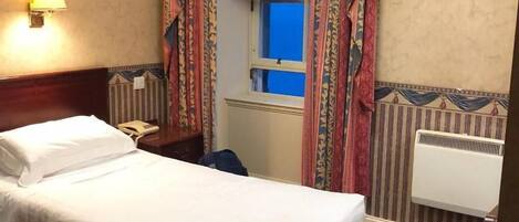 Una camera d'albergo con un letto, un comodino, una lampada, una finestra con tende, un termosifone e un quadro appeso al muro.