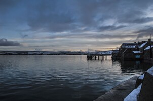 Una escena frente al agua con un muelle que se adentra en aguas tranquilas, rodeado de edificios con nieve en los tejados, lo que sugiere un clima frío.