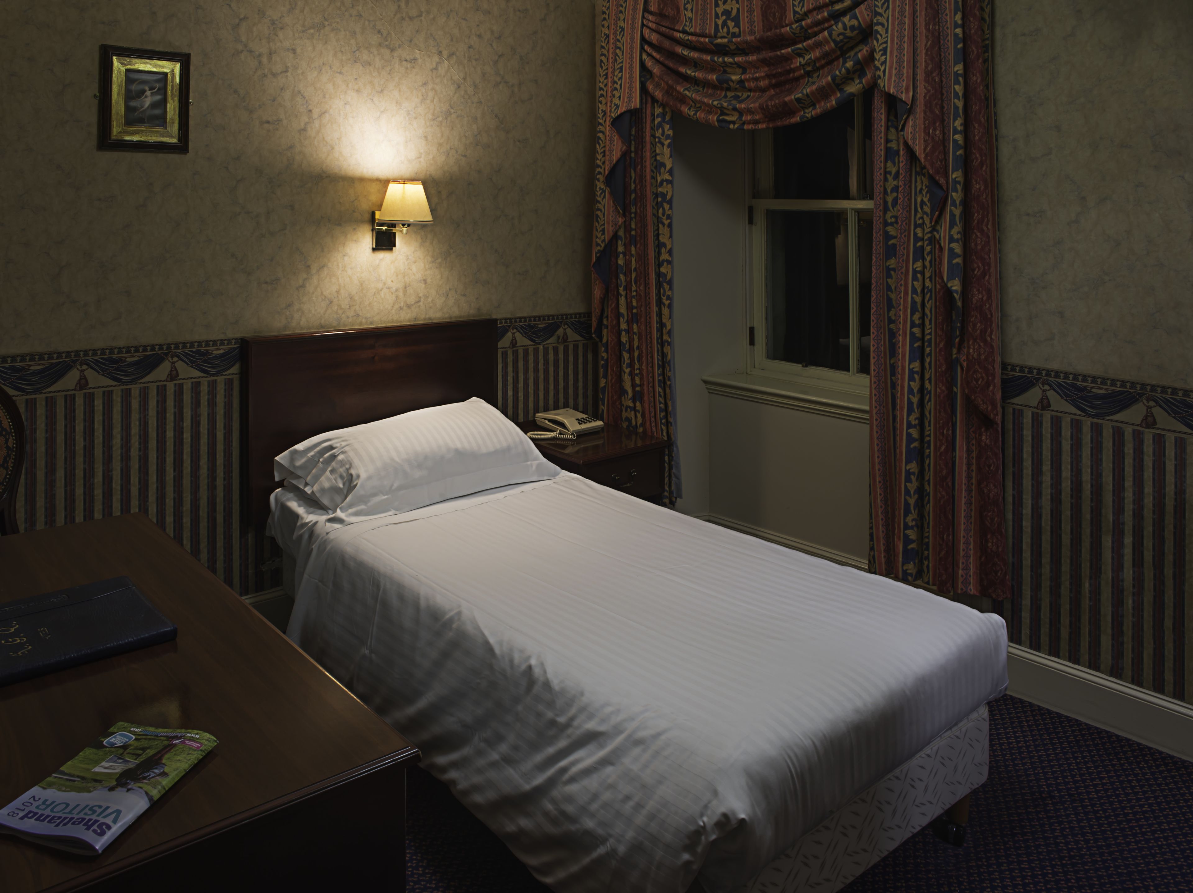 Une chambre d’hôtel avec un lit simple, une table de chevet avec une lampe, un téléphone et une fenêtre avec des rideaux.