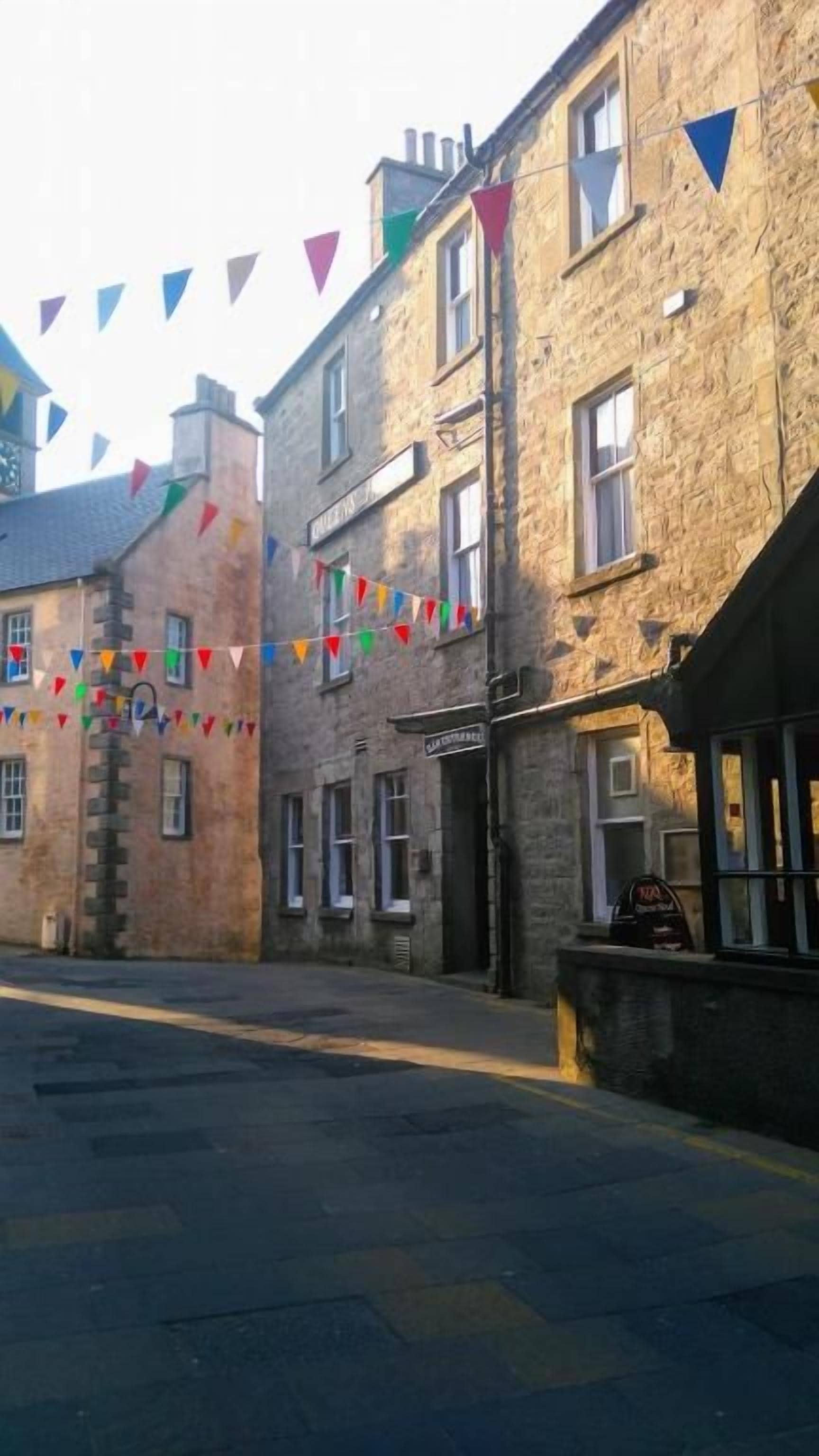 Une rue bordée d’immeubles en pierre, dont certains arborent des enseignes, sous un ciel dégagé.