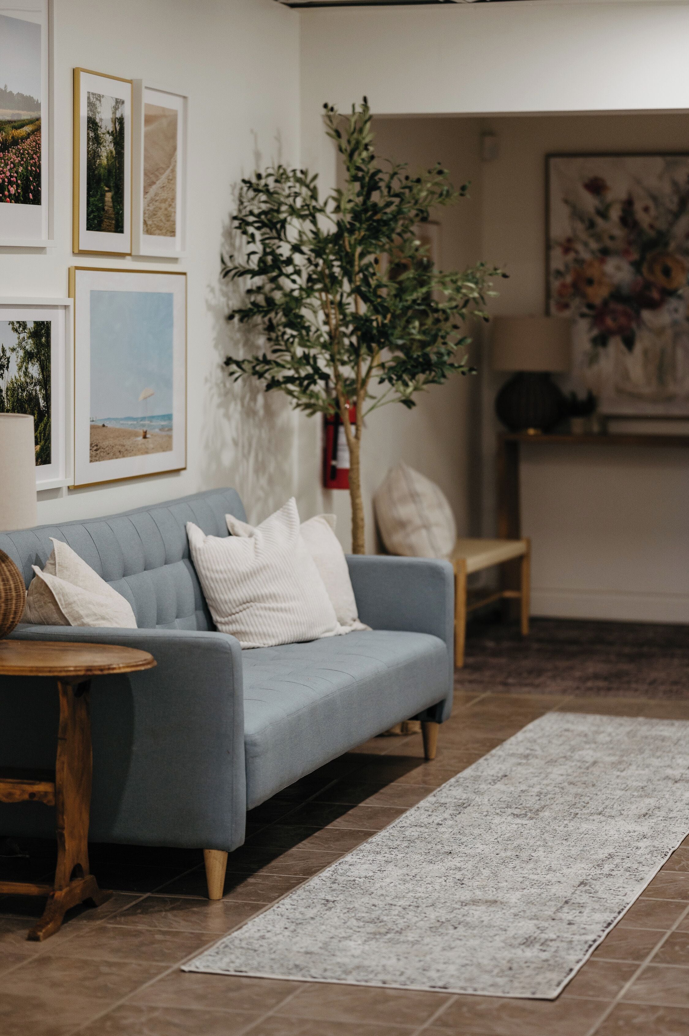 Un salon avec un canapé gris, une table d’appoint en bois, un tapis blanc et des tableaux encadrés accrochés au mur.