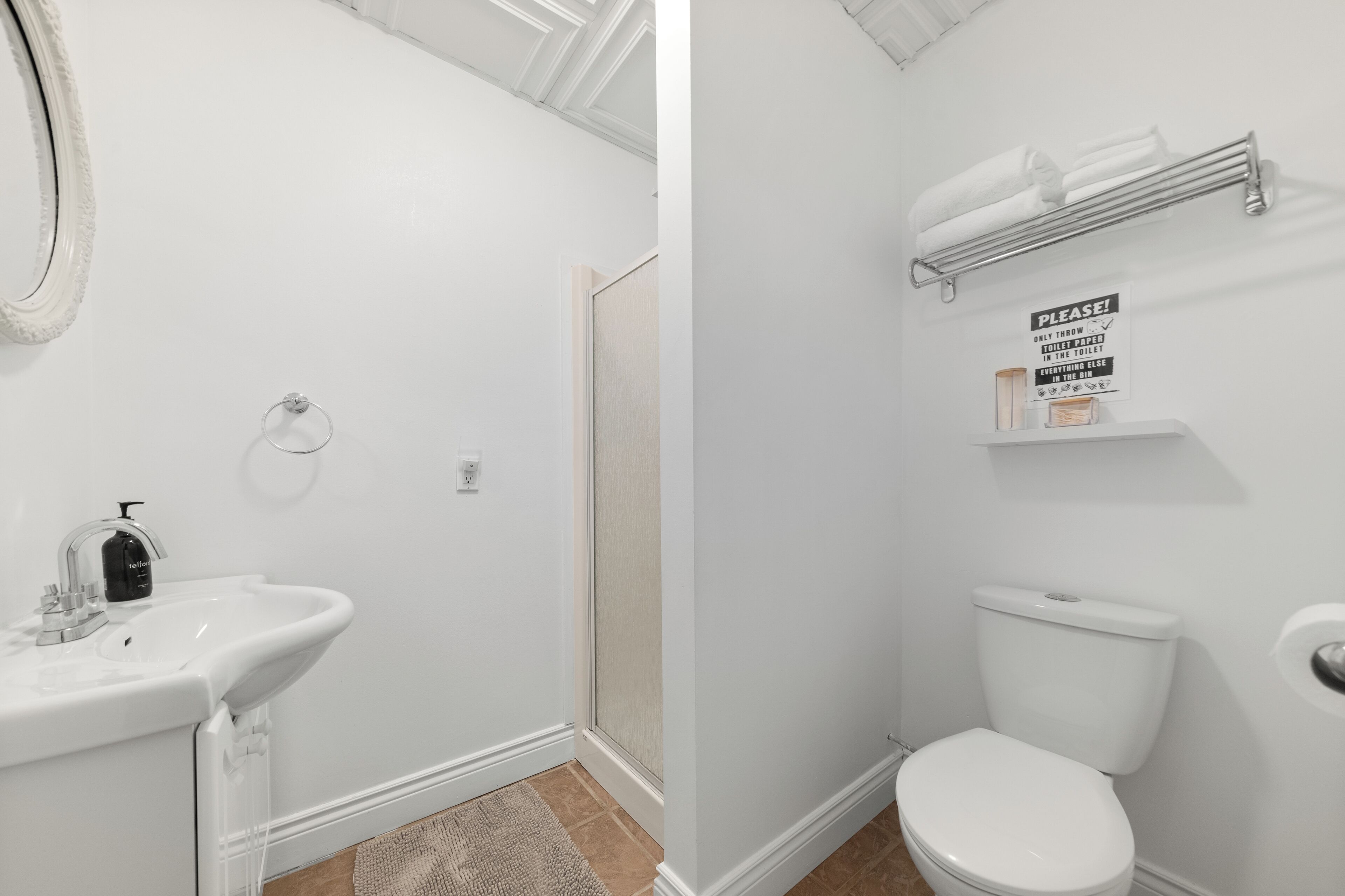 A bathroom with a white sink, toilet, and shower. There is a towel rack above the toilet and a sign on the wall.