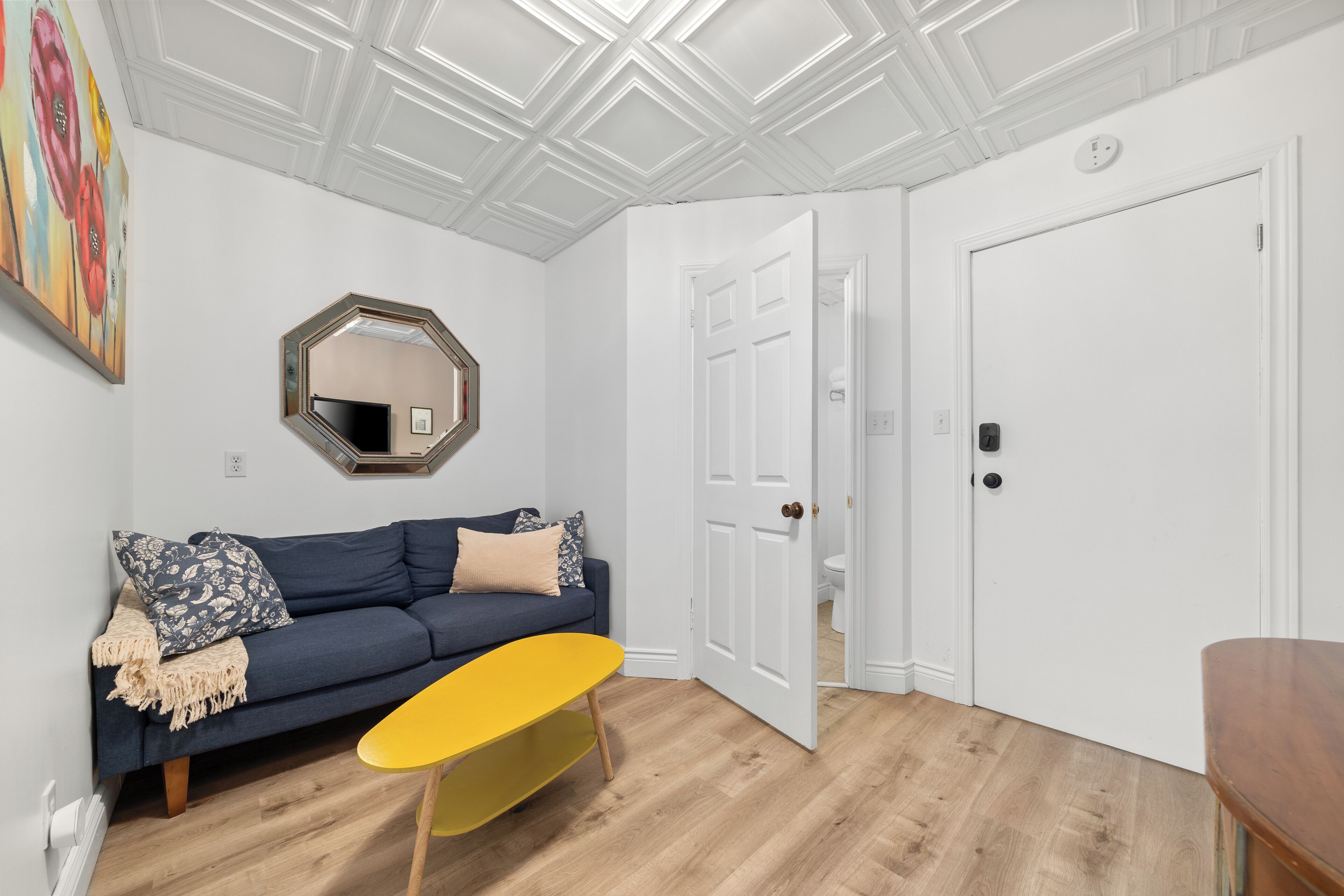 A room with a blue sofa, a yellow table, a white door, and a ceiling with a geometric pattern.