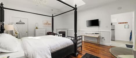 A four-poster bed with white linens, a chandelier, a flat-screen TV, and a desk with a chair in a modern bedroom.