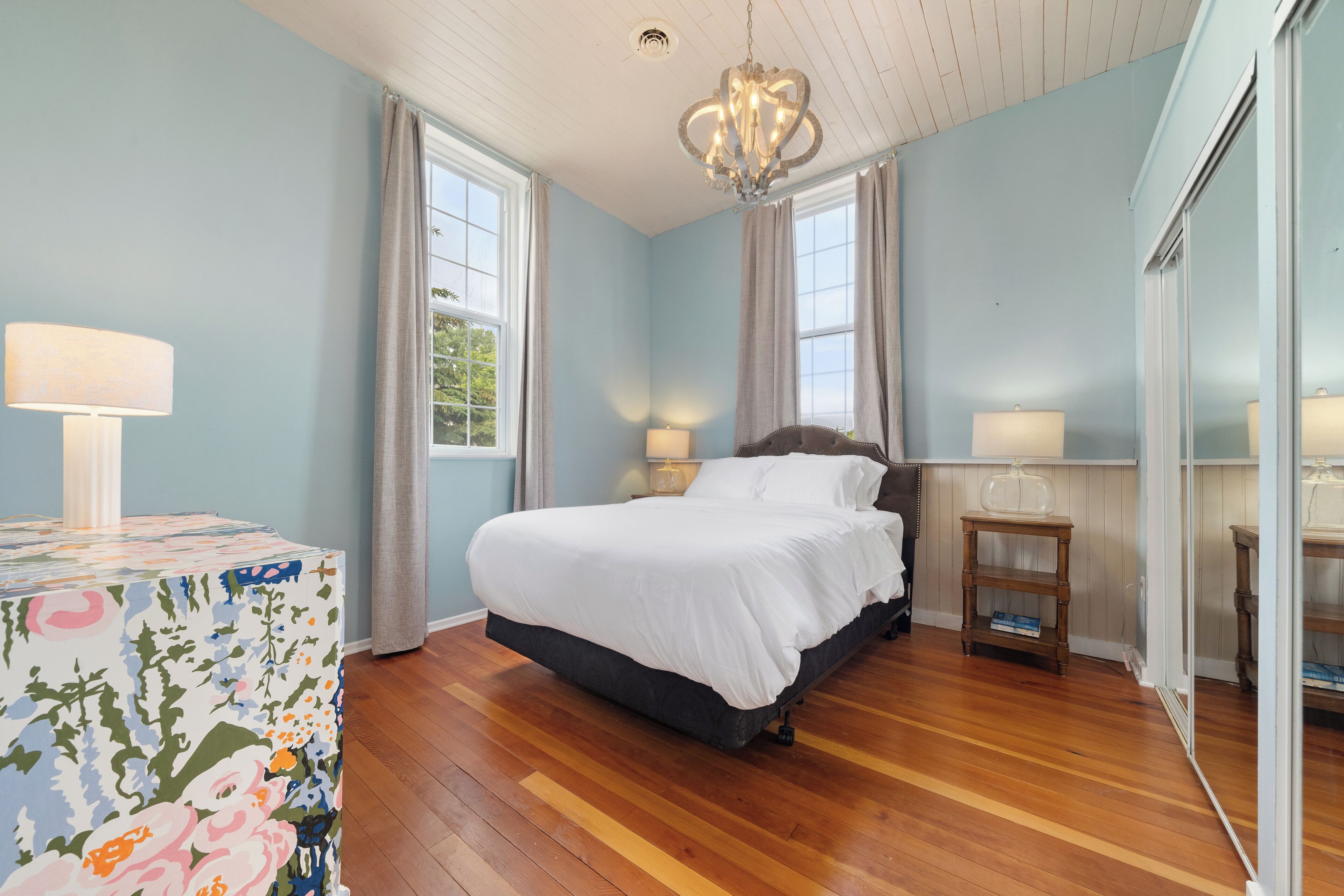 A bedroom with a large bed, two bedside lamps, a chandelier, and a floral-patterned table.