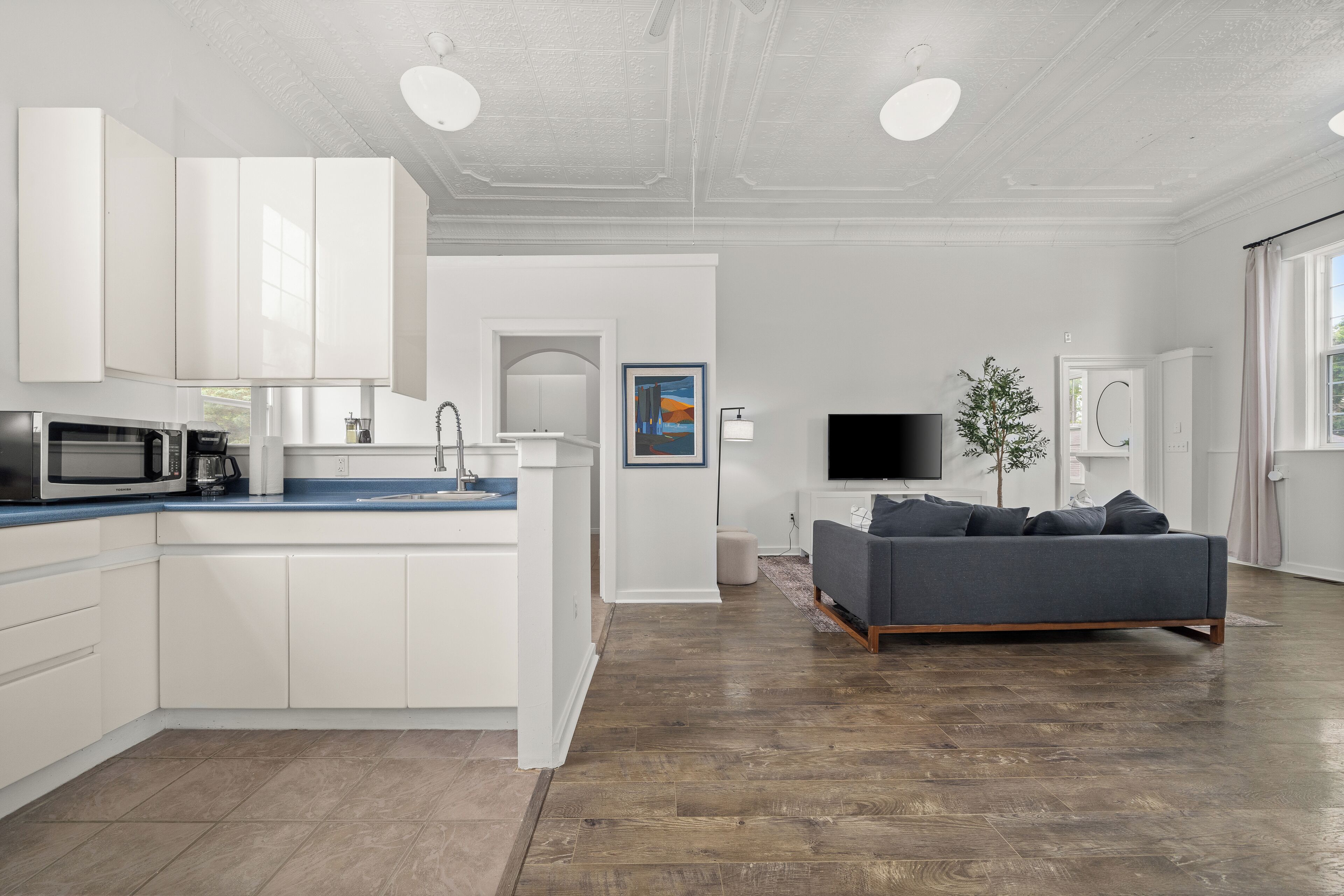Eine moderne Küche mit weißen Schränken, integriertem Mikrowellenherd und einer Kaffeemaschine. Ein Wohnbereich mit grauem Sofa, Flachbildfernseher und einer kleinen Pflanze.