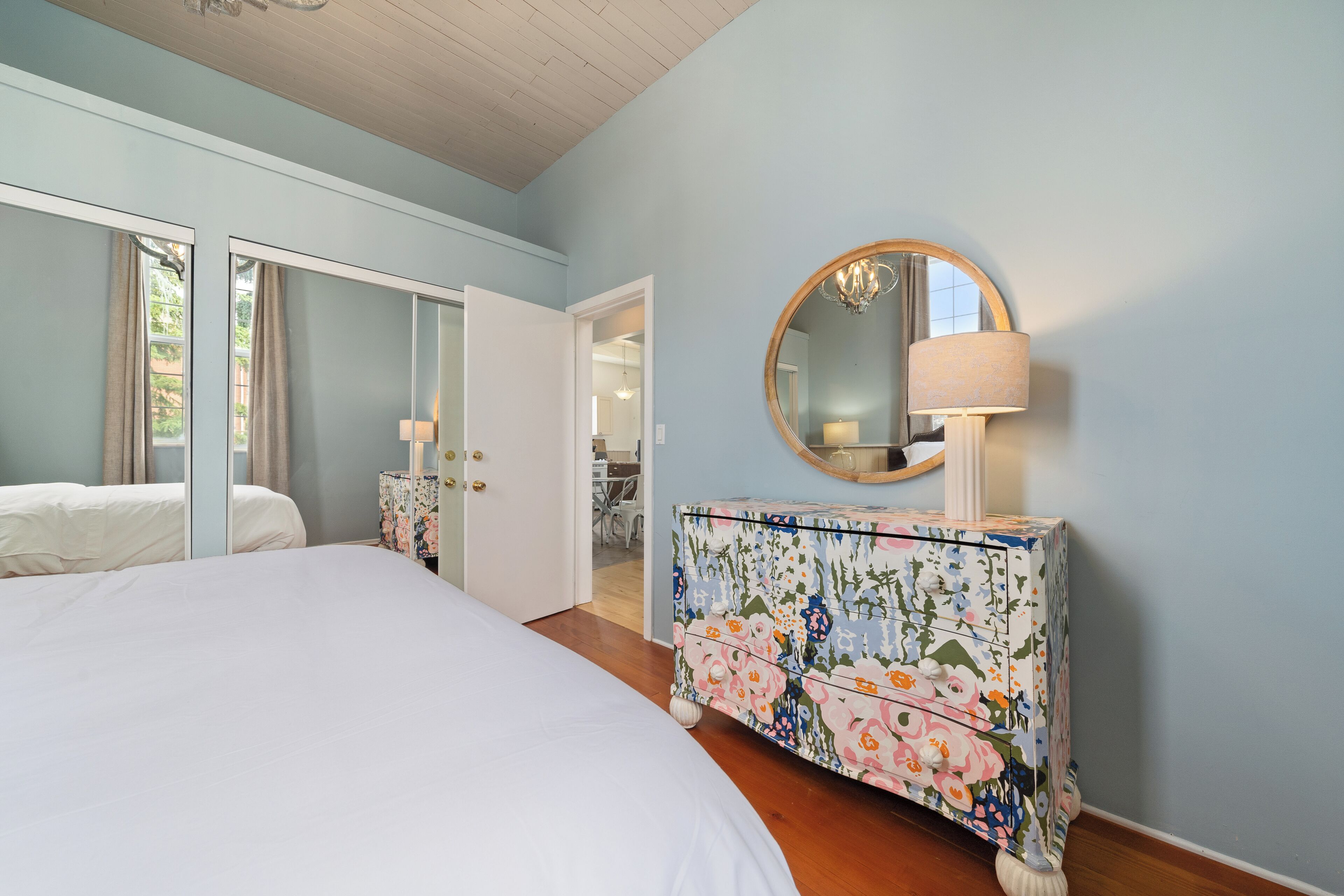 A bedroom with a bed, a dresser with floral patterns, a round mirror, and a door leading to another room.