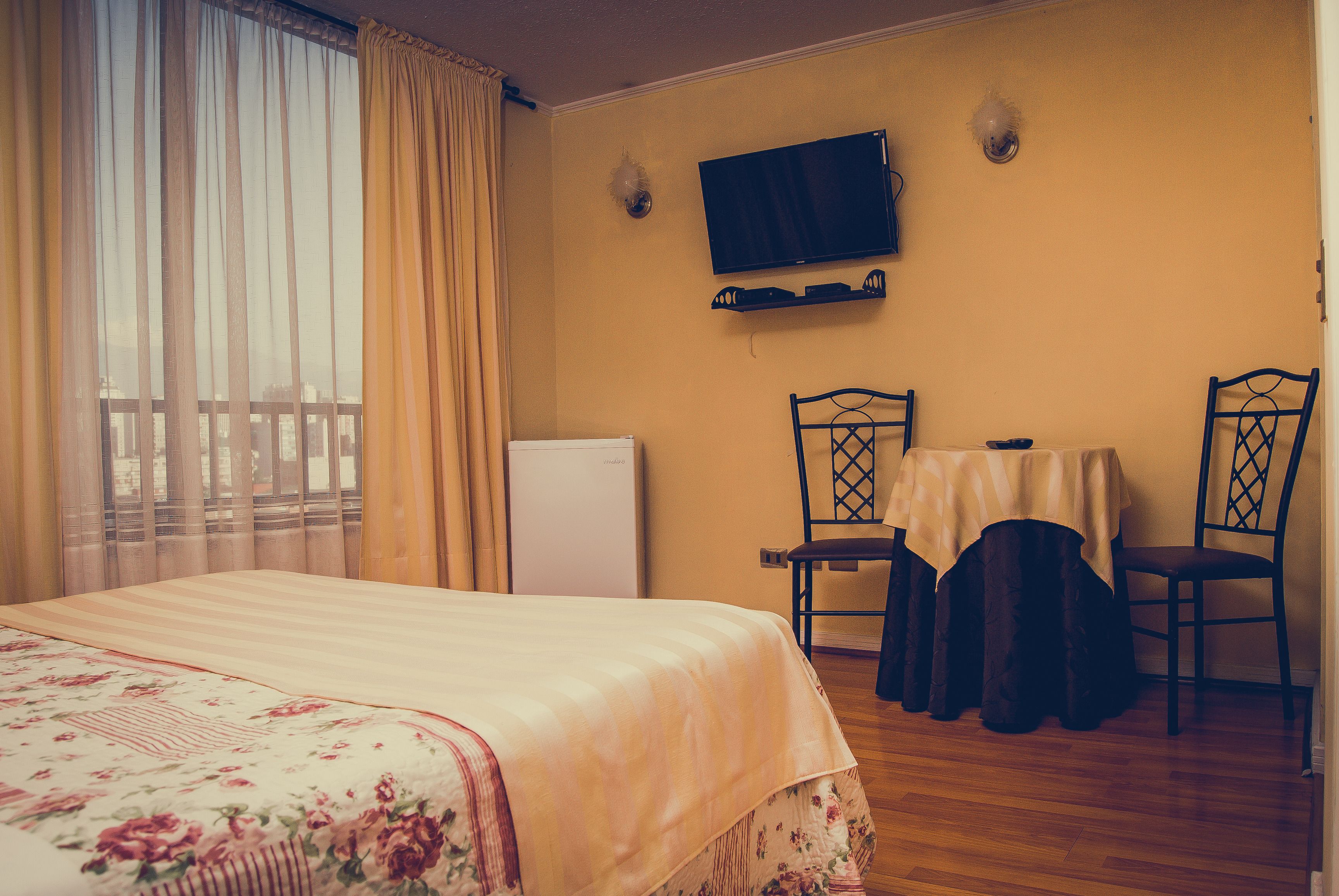 Habitación de hotel con cama, una mesa con sillas, televisor y ventana con cortinas.
