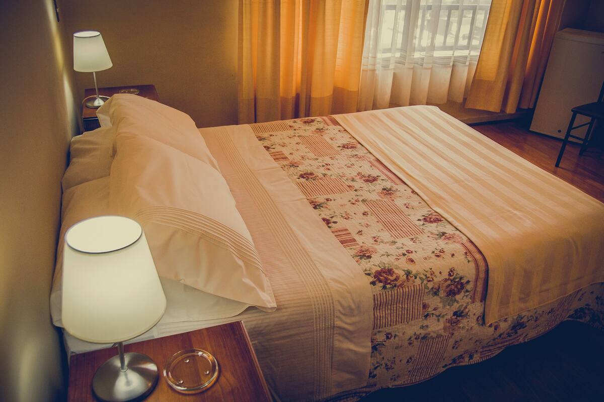 Un lit bien fait, recouvert d’une couvre-lit à motifs floraux, avec une lampe de chevet et une table de nuit en bois.