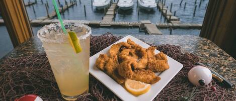 A table with a plate of fried seafood, a margarita with a lime wedge, and a view of boats on water.