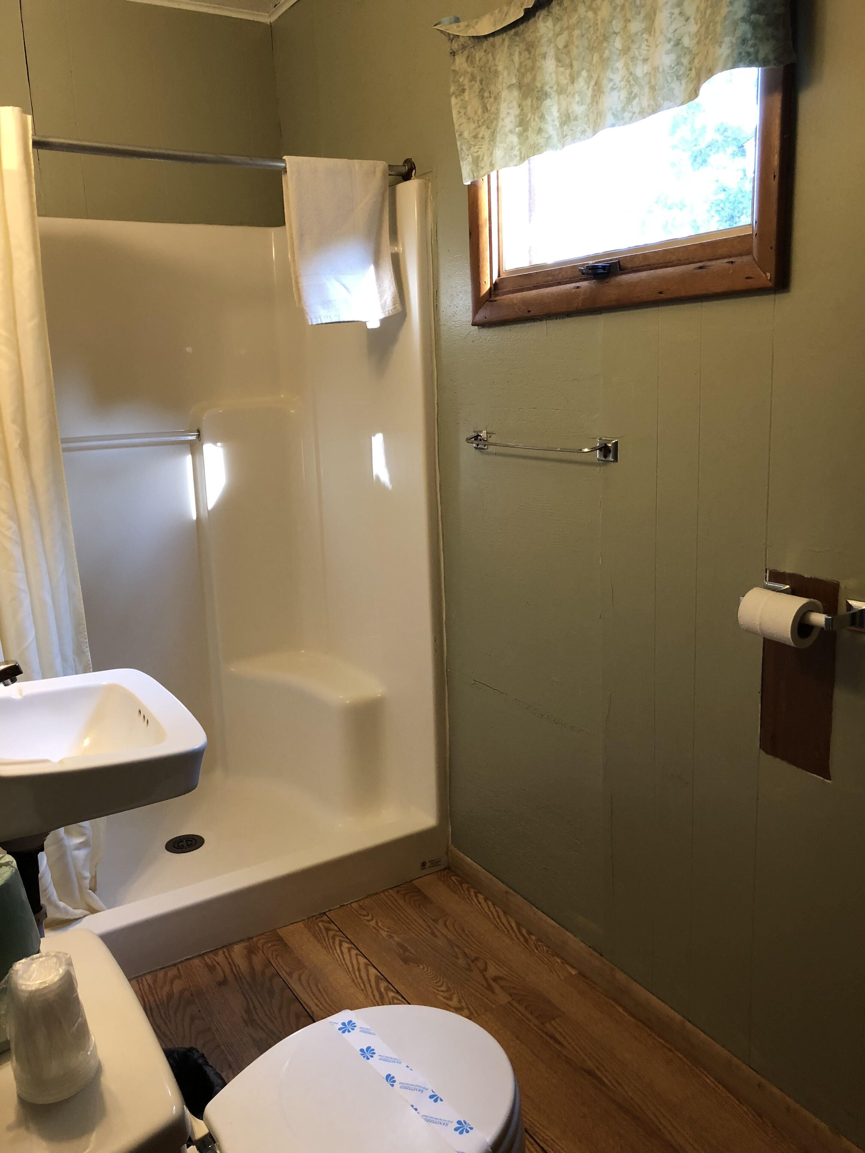 A bathroom with a shower, toilet, sink, and a window with a curtain.