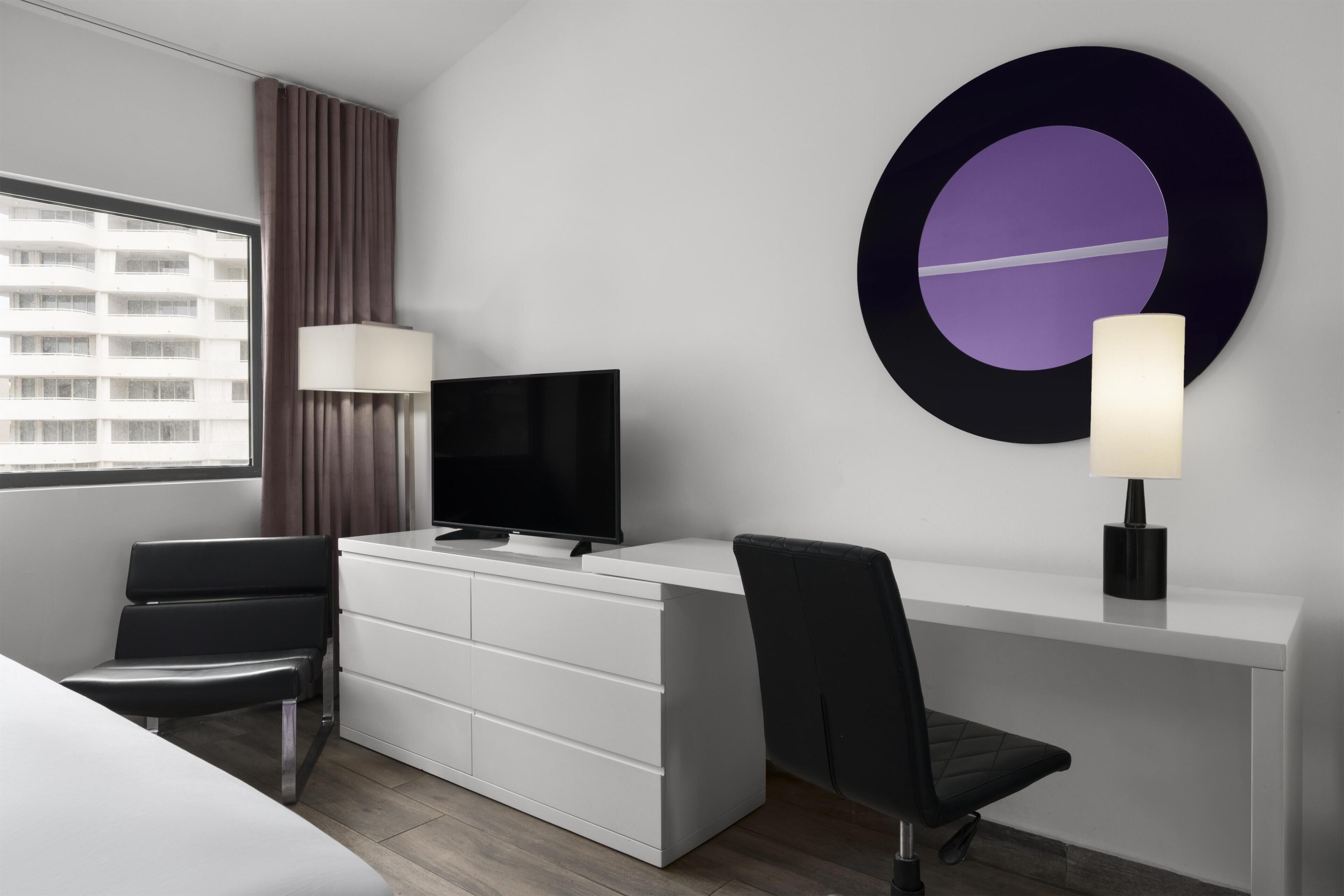 Habitación de hotel moderna con televisor de pantalla plana, escritorio con silla, ventana grande con cortinas y una obra de arte circular montada en la pared.