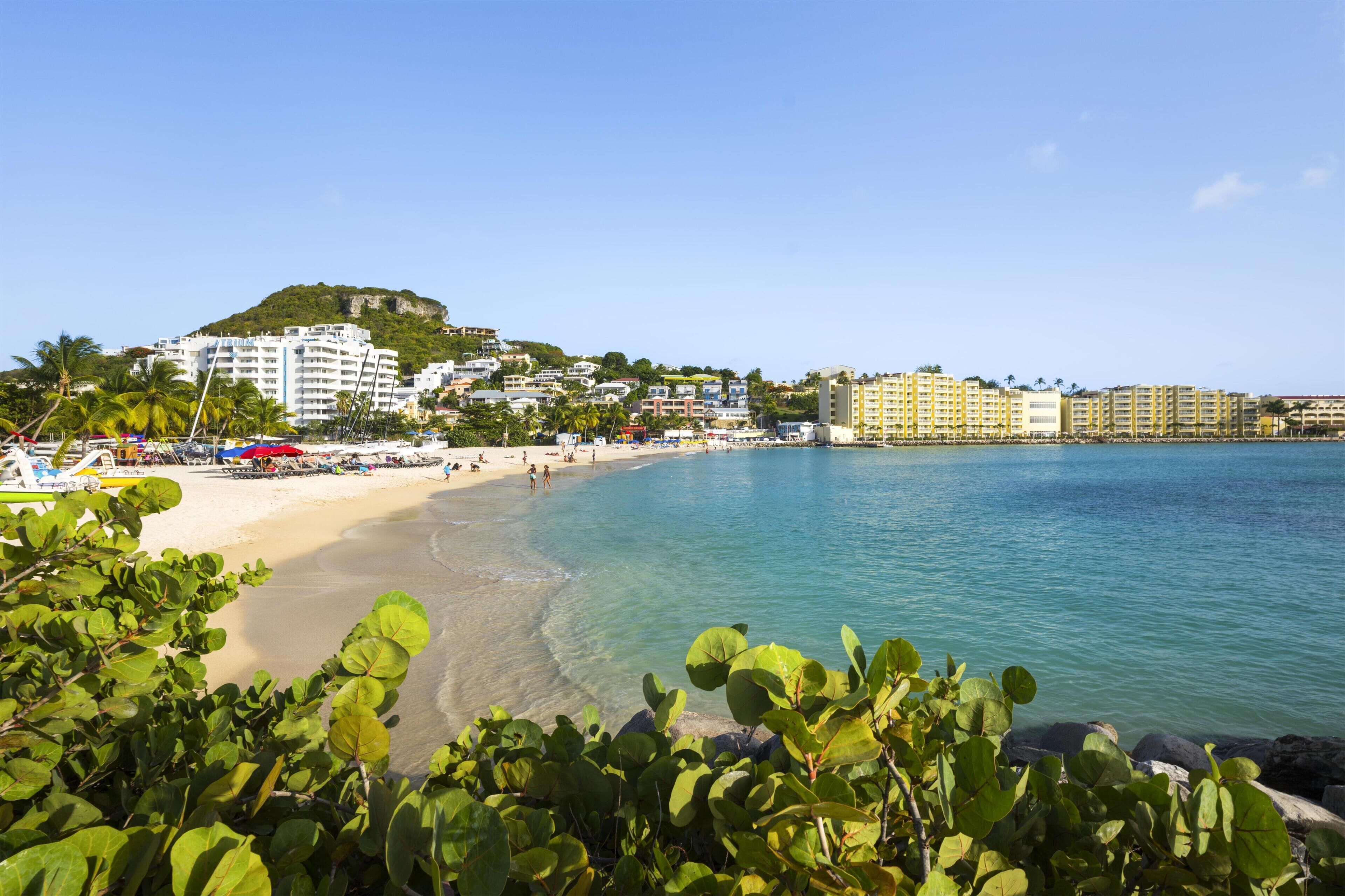 A coastal town with a beach, clear blue water, and buildings in the background.