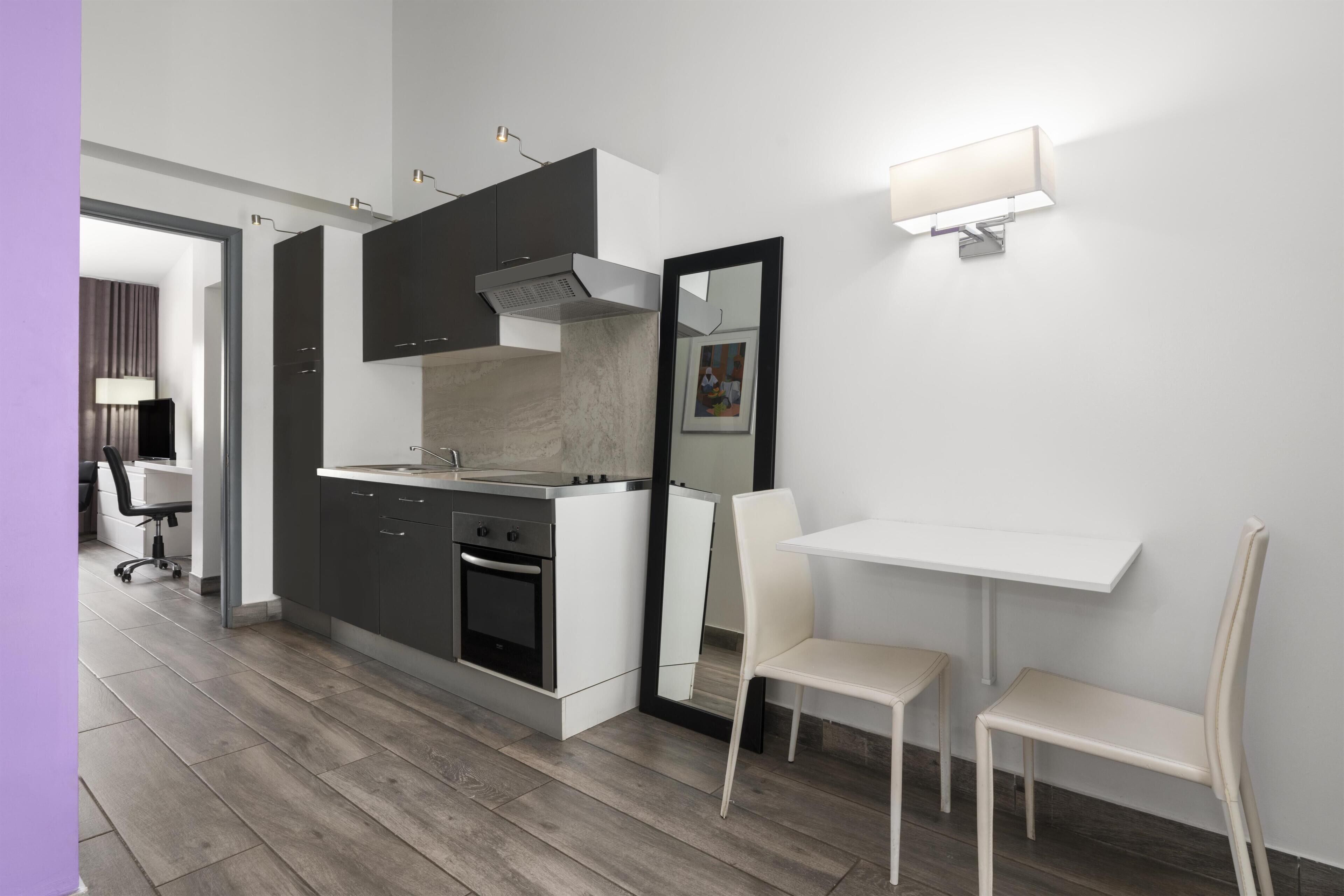 Una cocina compacta con electrodomésticos modernos, un comedor con mesa y sillas blancas, y un espejo en la pared.