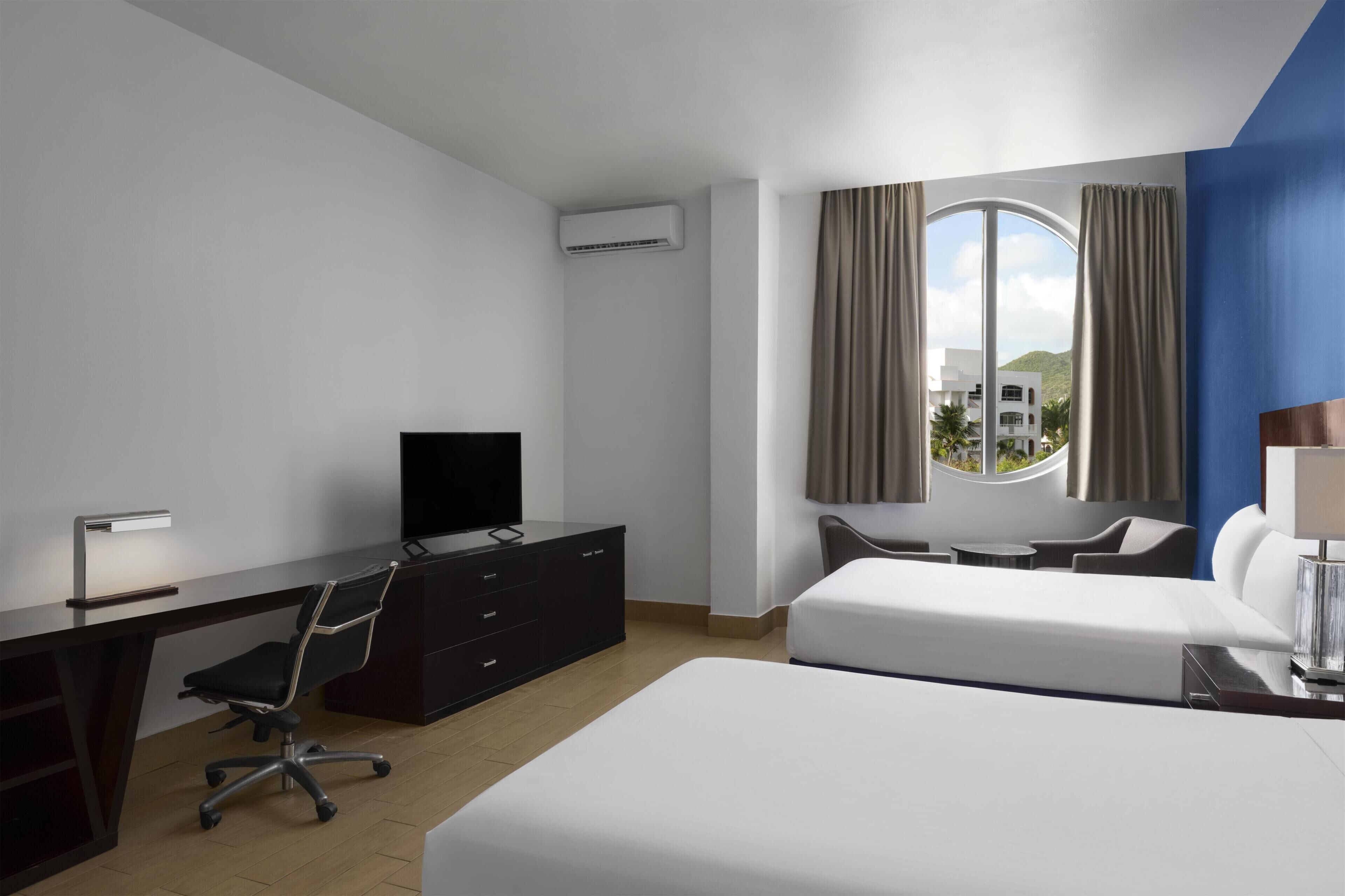 Habitación de hotel con cama, escritorio, silla, televisor y ventana con cortinas.