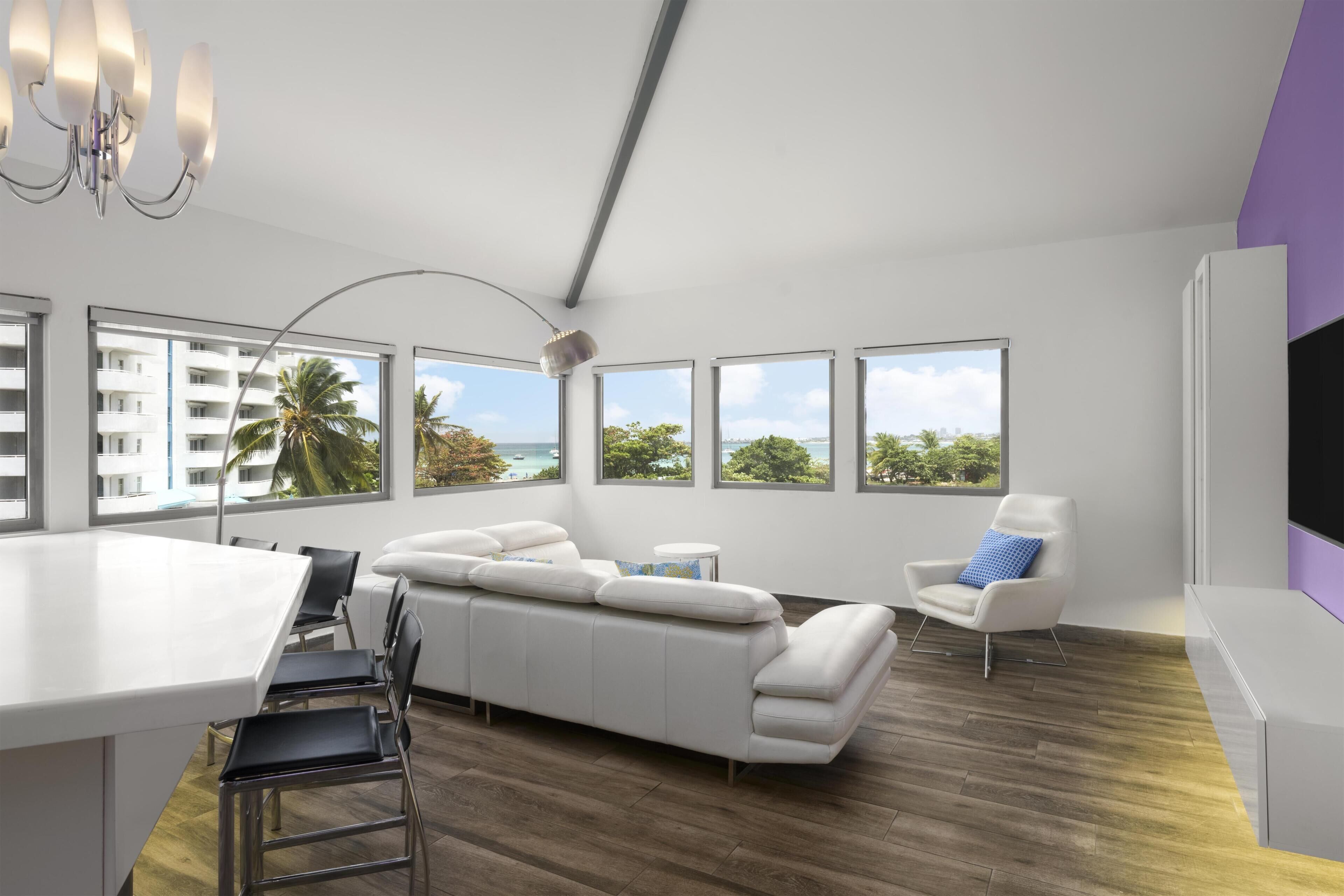 A modern living room with a sofa, a dining table, and a chair. There are large windows with a view of the sea and buildings.