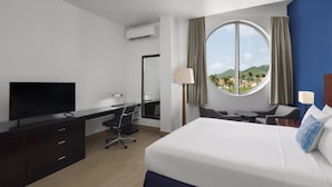 A hotel room with a bed, desk, chair, television, and a large window with a view of a landscape. - Commodore Suites, SureStay Collection by Best Western (Simpson Bay)