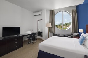 A hotel room with a bed, desk, chair, television, and a large window with a view of a landscape. - Commodore Suites, SureStay Collection by Best Western (Simpson Bay)