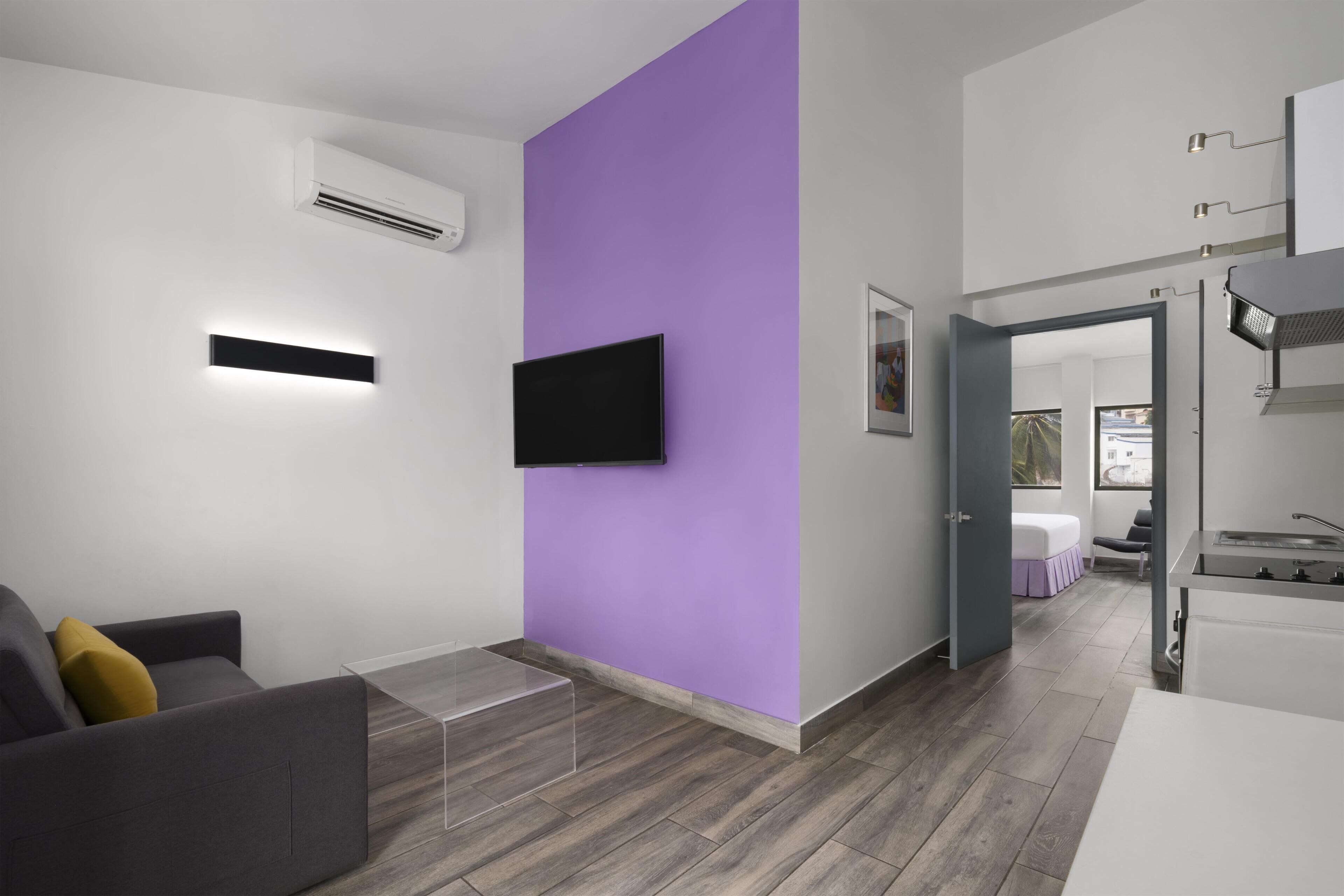 Quarto de hotel moderno com televisão de tela plana fixada numa parede roxa, uma mesa pequena, um sofá e uma área de cozinha compacta.