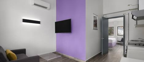A modern hotel room with a flat-screen TV mounted on a purple wall, a small table, a sofa, and a kitchenette area.