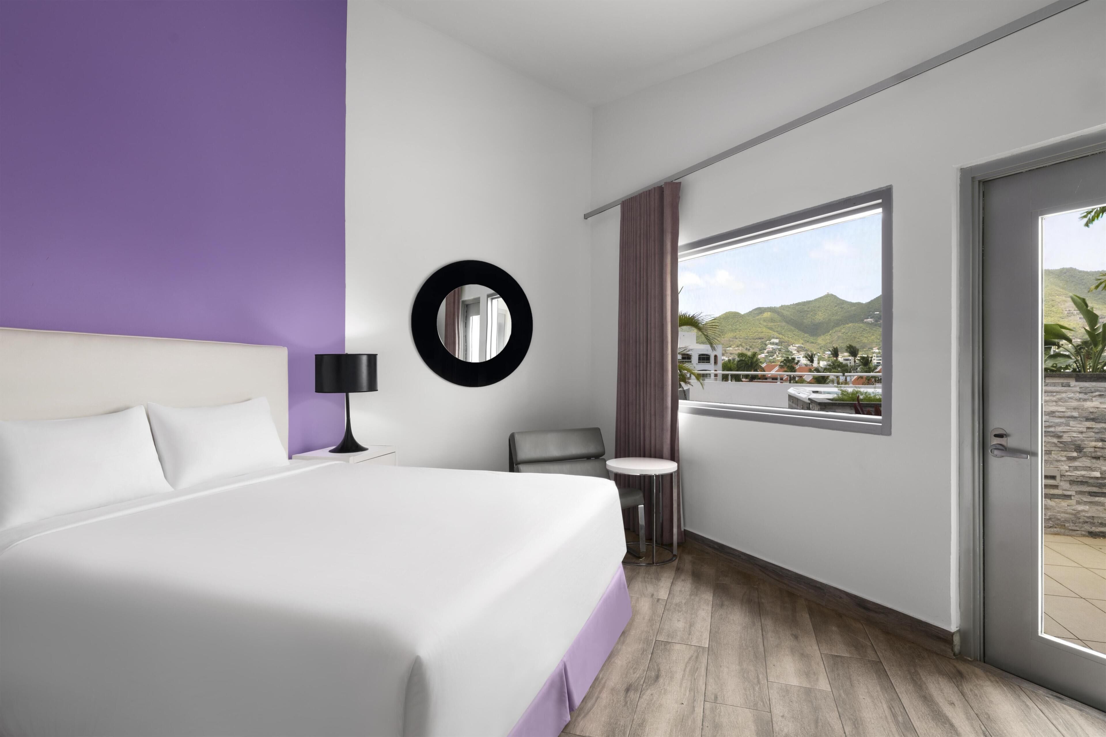 A hotel room with a bed, a round mirror, a small table, and a view of a mountainous landscape through a large window.