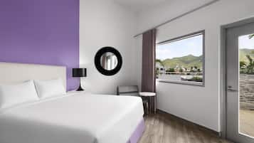 A hotel room with a bed, a round mirror, a small table, and a view of a mountainous landscape through a large window.