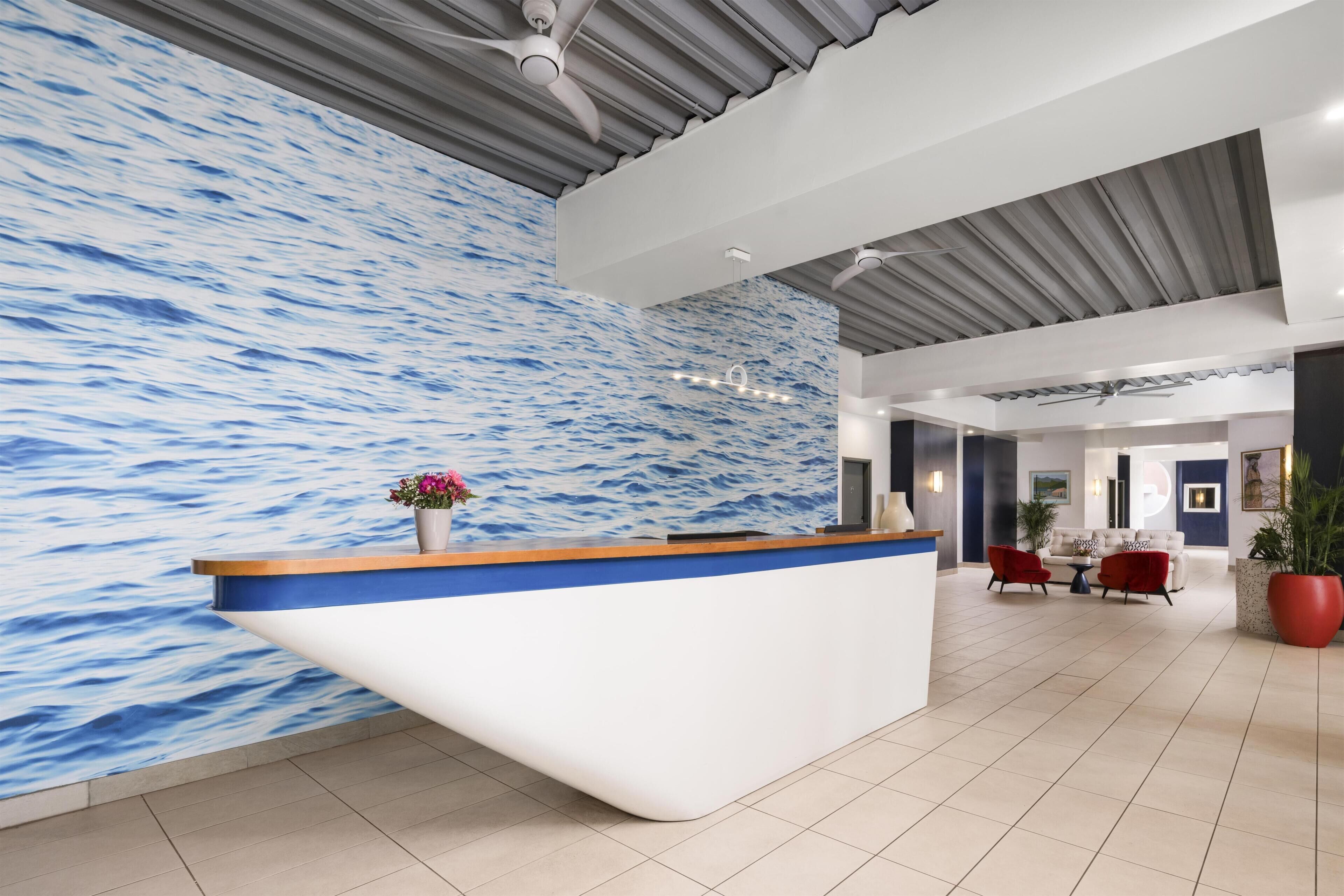Un mostrador de recepción con un muro detrás que presenta un diseño de olas azules y blancas, un ventilador de techo y una zona de estar con sillas rojas y una mesa.