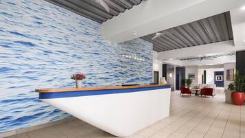 Un mostrador de recepción con un muro detrás que presenta un diseño de olas azules y blancas, un ventilador de techo y una zona de estar con sillas rojas y una mesa.