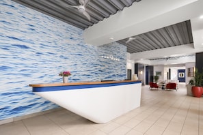 Een receptiebalie met een muur achter zich met een blauw-wit golfpatroon, een plafondventilator en een zithoek met rode stoelen en een tafel.