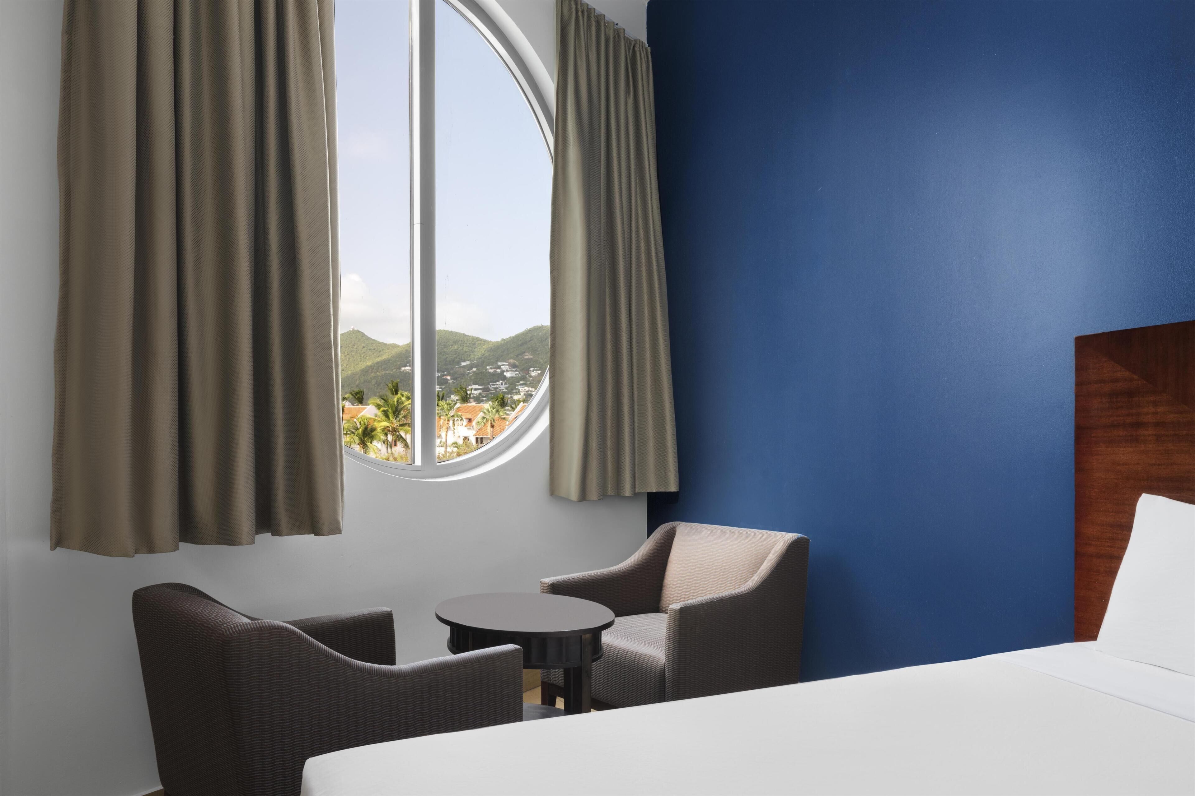 Habitación de hotel con cama, dos sillas, una mesita y un ventanal con cortinas.