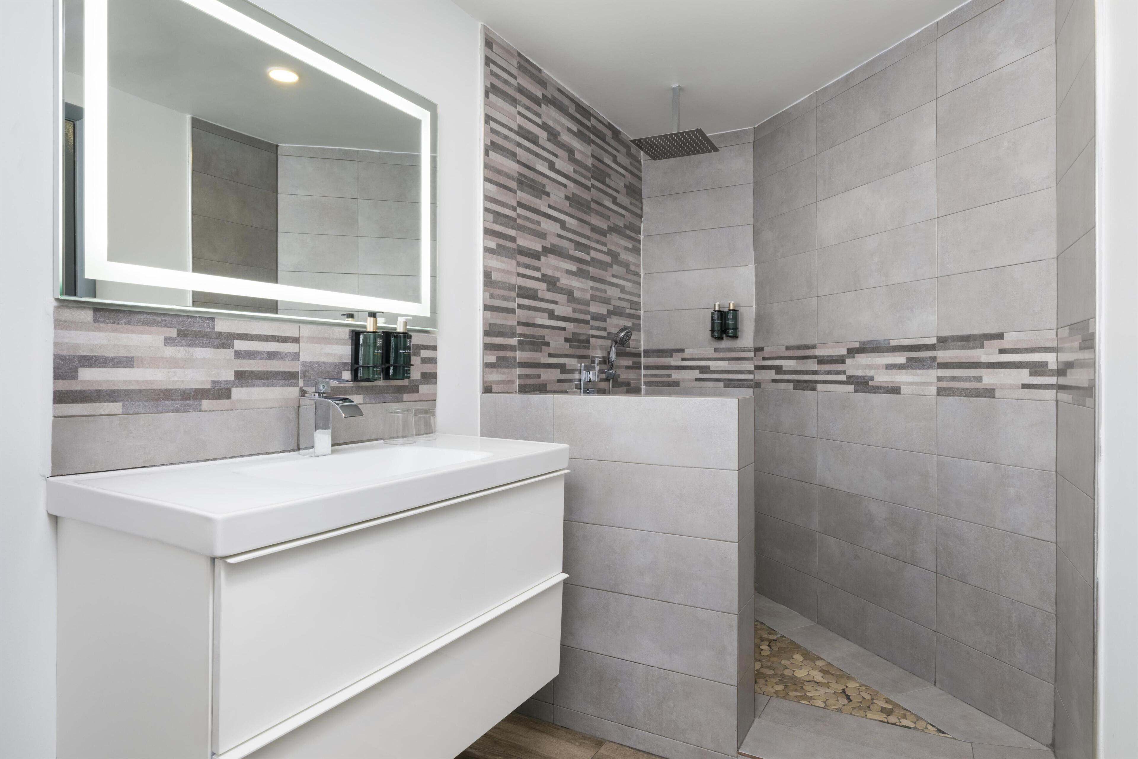Un baño moderno con tocador blanco, un espejo grande y zona de ducha con azulejos grises.