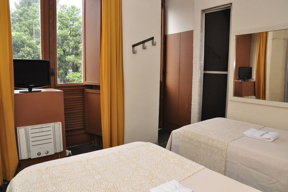 A hotel room with two beds, a television, a window with curtains, and a mirror.