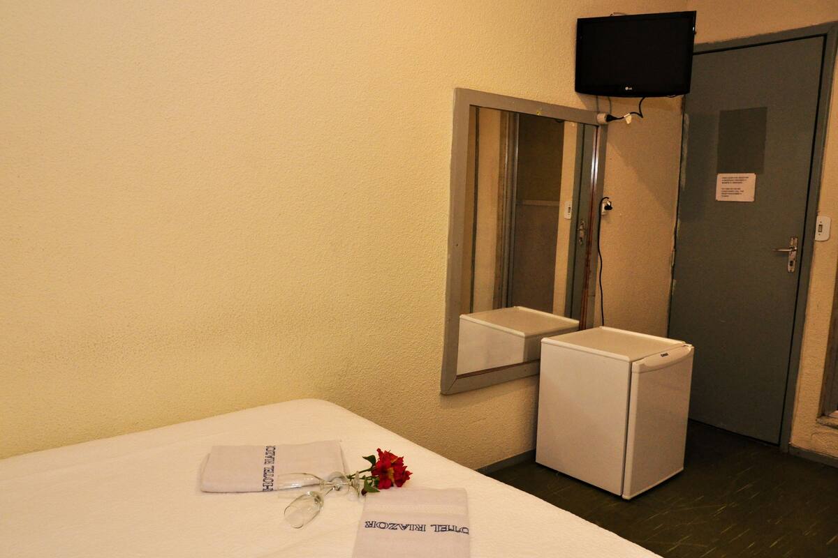 A hotel room with a bed, a small white appliance, a mirror, and a television mounted on the wall.