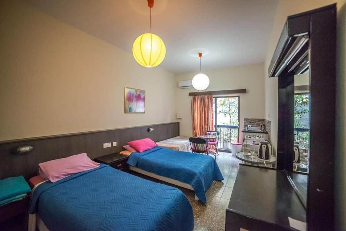 Quarto de hotel com duas camas, uma escrivaninha, uma cadeira, uma mesa com chaleira e uma janela com cortinas.
