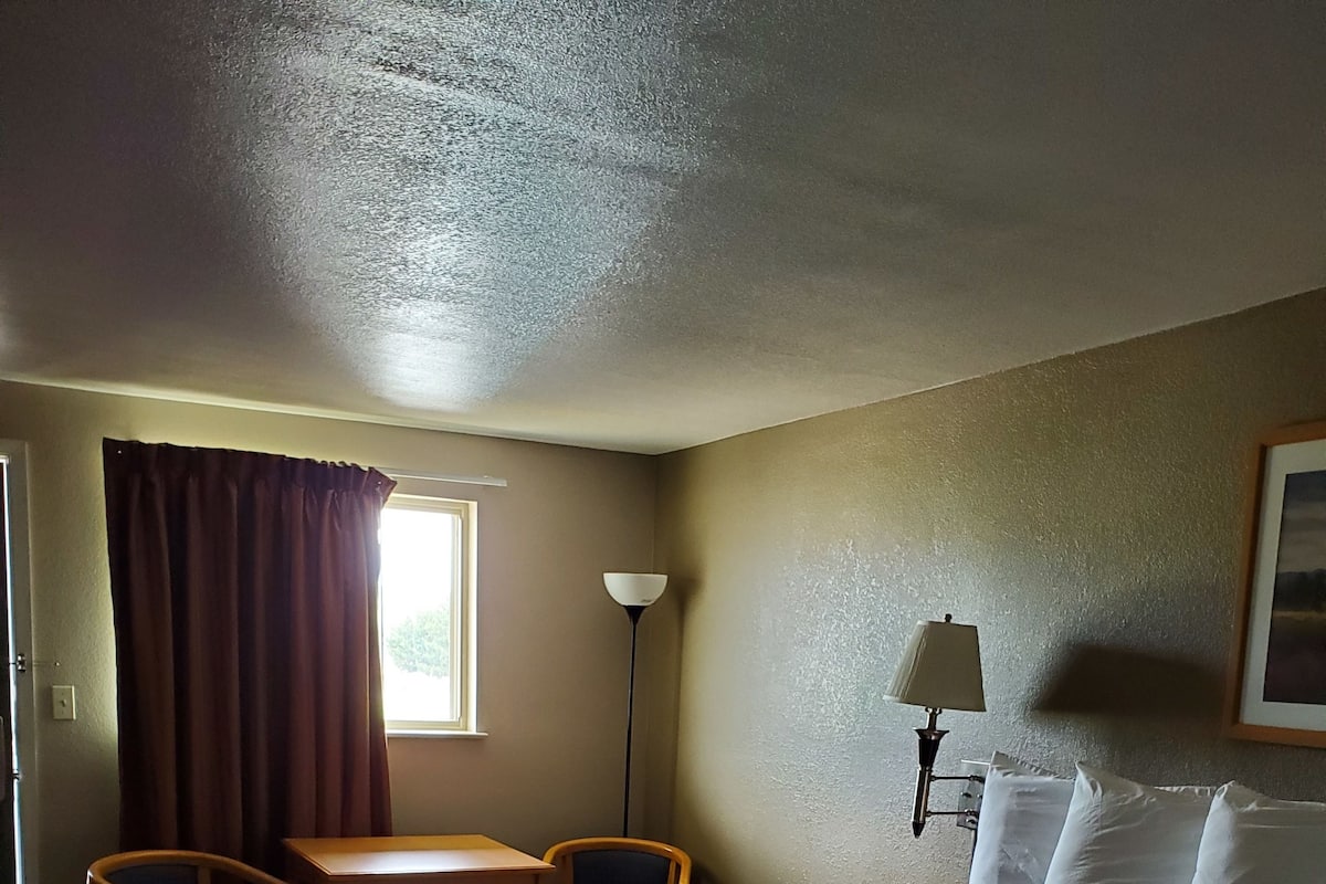 Quarto de hotel com cama, escrivaninha, duas cadeiras, luminária e janela com cortinas.