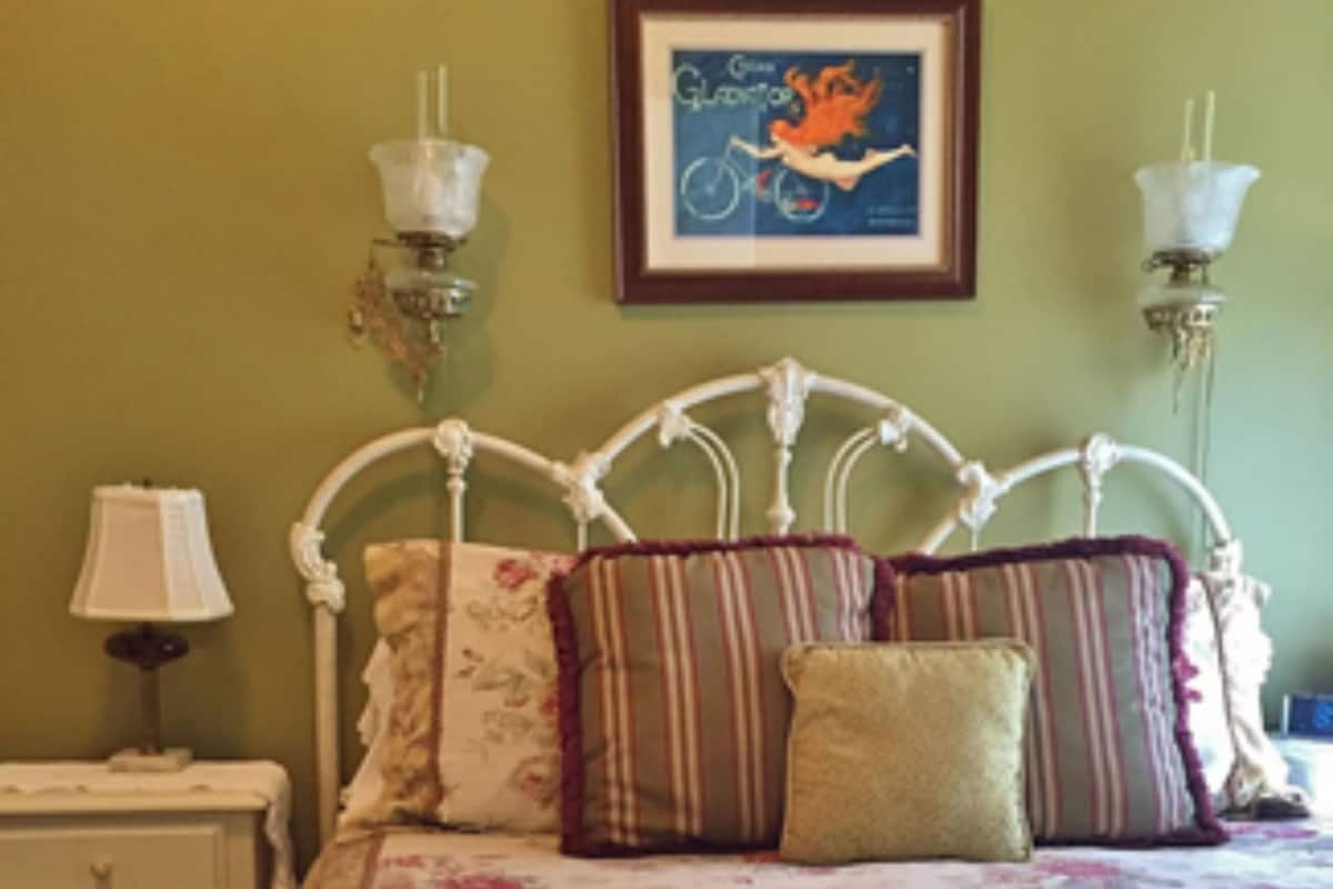 A bed with a floral bedspread, a white metal headboard, and multiple pillows. There is a framed picture on the wall above the bed.