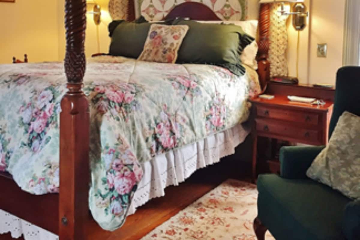 A four-poster bed with floral bedding, a green armchair, and a patterned wall hanging.