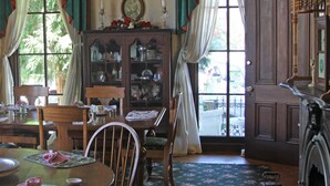 A dining room with a wooden table, chairs, a cabinet with glass doors, and a large window with curtains. - Inn At The Canal Bed & Breakfast (Chesapeake City)