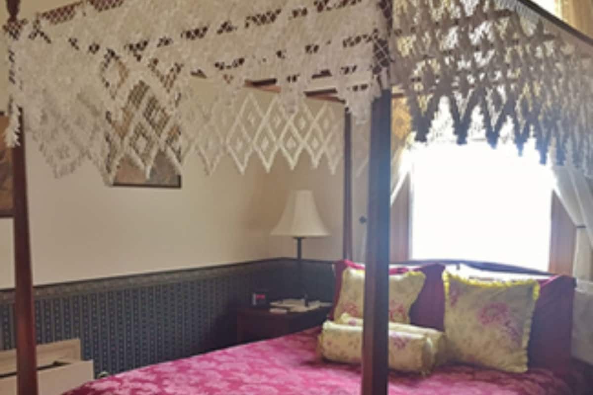 A four-poster bed with a floral bedspread, a nightstand with a lamp, and a patterned wall.