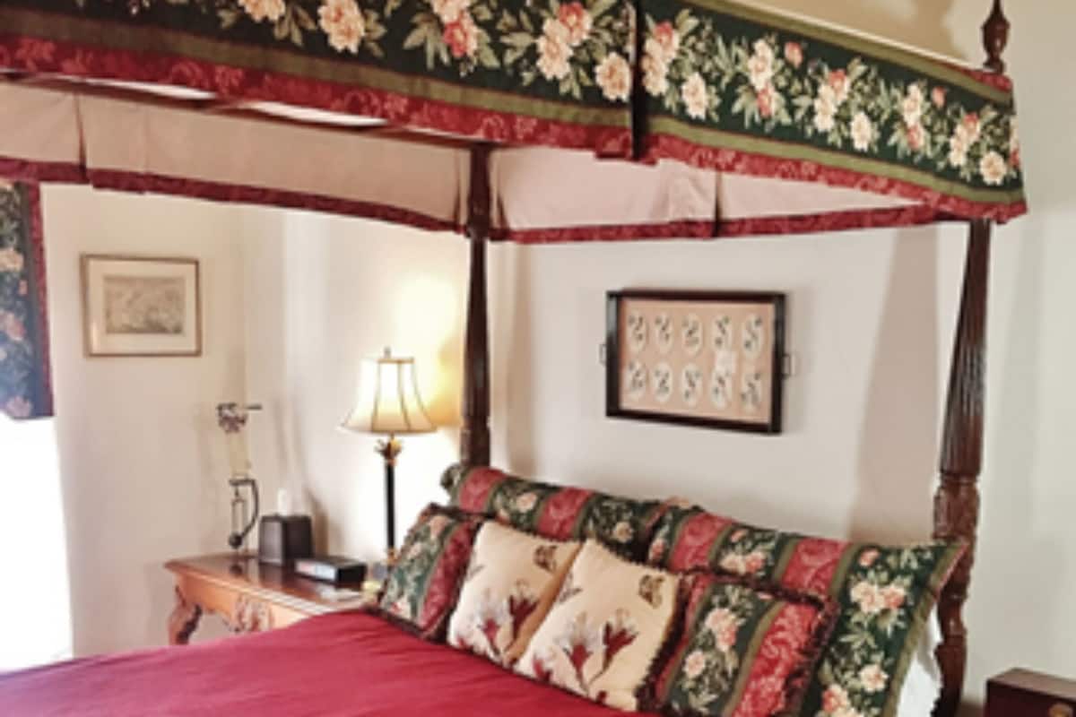 A four-poster bed with floral bedspread and pillows, a bedside table with a lamp, and a framed picture on the wall.