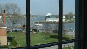 A view through a window of a river with a large ship docked at a pier, a sailboat, and a small building with a green roof. - Inn At The Canal Bed & Breakfast (Chesapeake City)