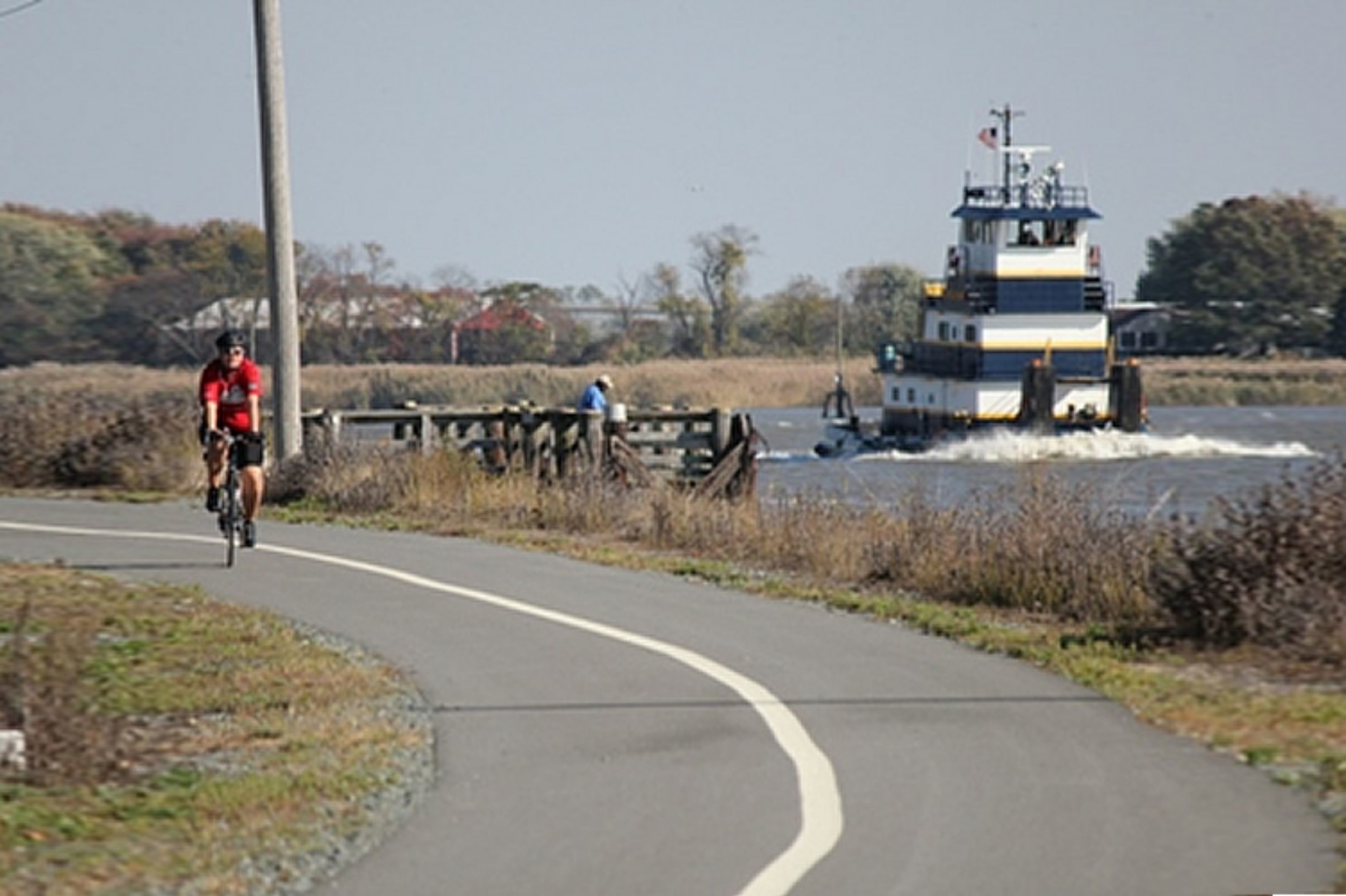 A cyclist on a paved path beside a body of water with a boat in the background.
