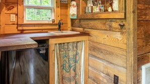 A rustic wooden kitchen with a built-in dishwasher, a window with a view of trees, and shelves with jars and kitchen utensils. - The Owl's Perch (5 inch clearance needed) (Robbinsville)