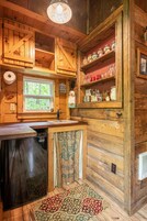 Una cocina rústica de madera con lavavajillas incorporado, una ventana con vistas a árboles y estantes con frascos y utensilios de cocina.