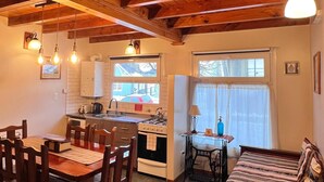 A cozy cabin interior with a wooden dining table, chairs, a sofa, a kitchen area with a stove and sink, and a window with curtains. - La Posta Apart (Ushuaia)