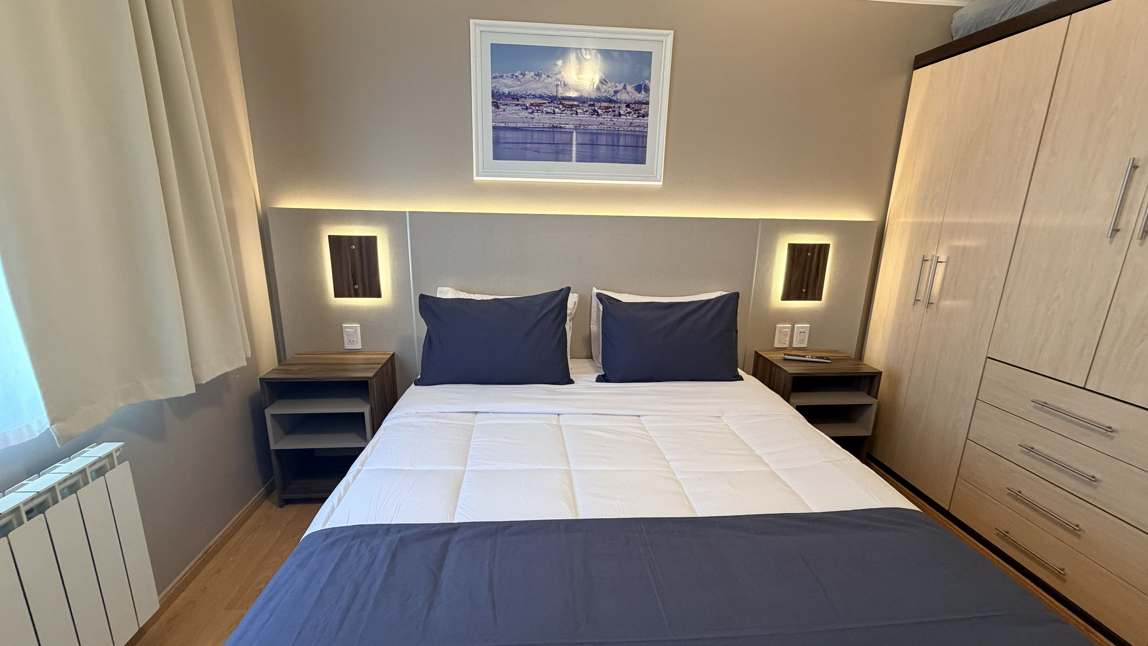 A hotel room with a large bed, two bedside tables, a wardrobe, and a framed picture on the wall.