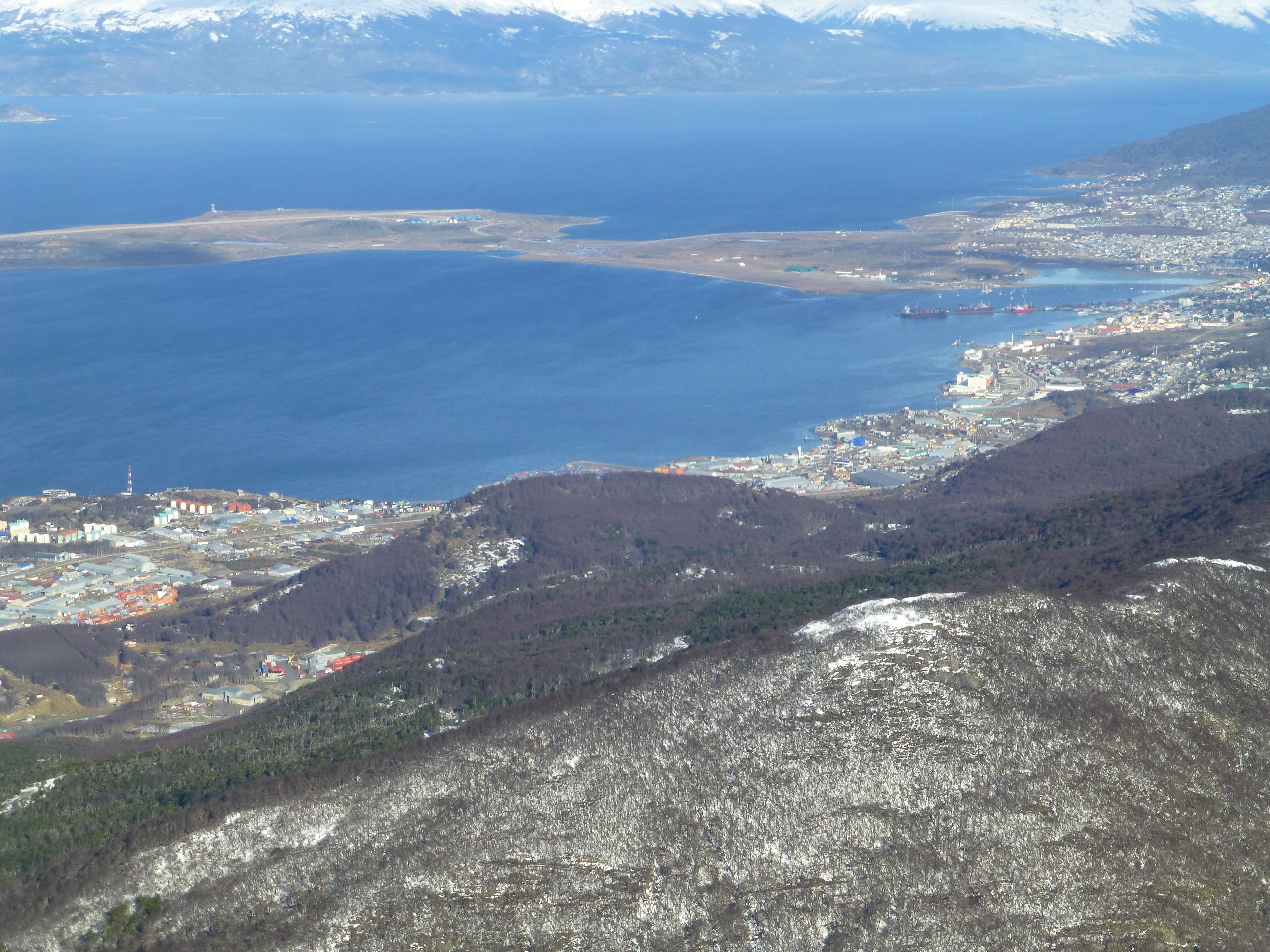 A coastal town with a mix of residential and industrial buildings, a large body of water, and snow-capped mountains in the background.